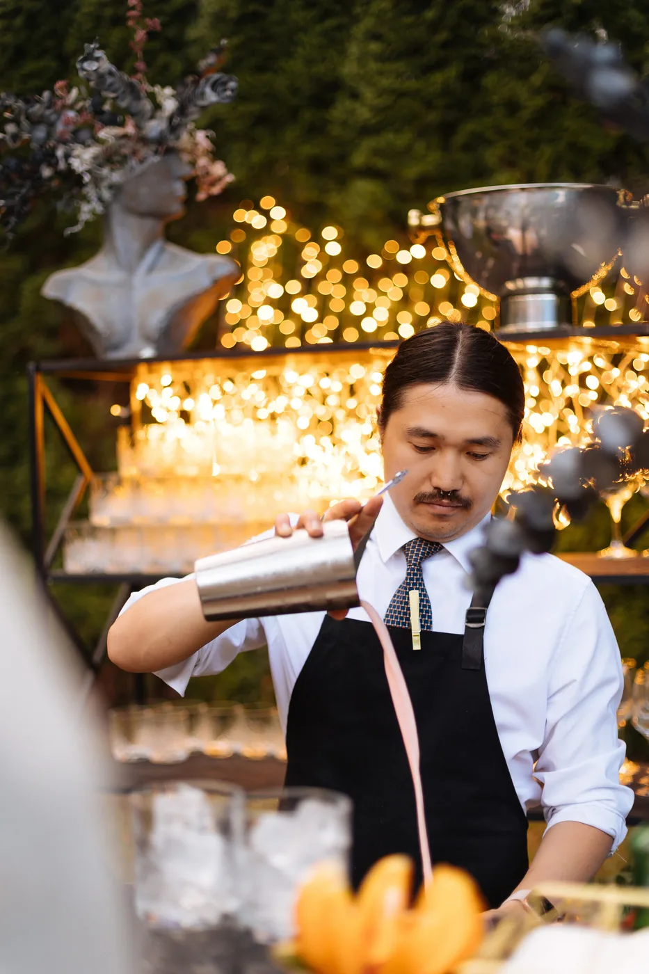 A bartender making a signature cocktail at a bar with string lighting