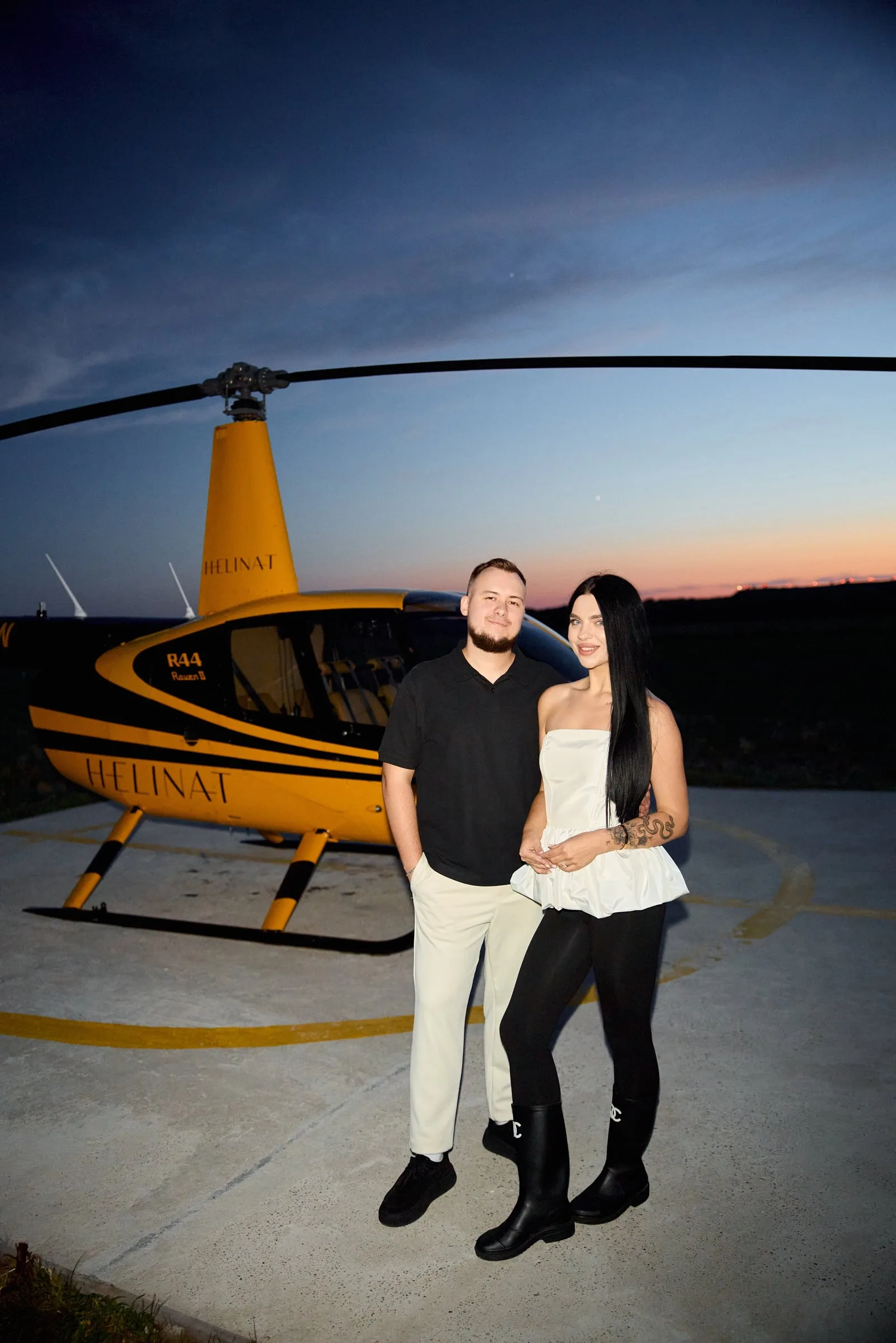 A couple standing on a helipad beside a yellow helicopter at dusk