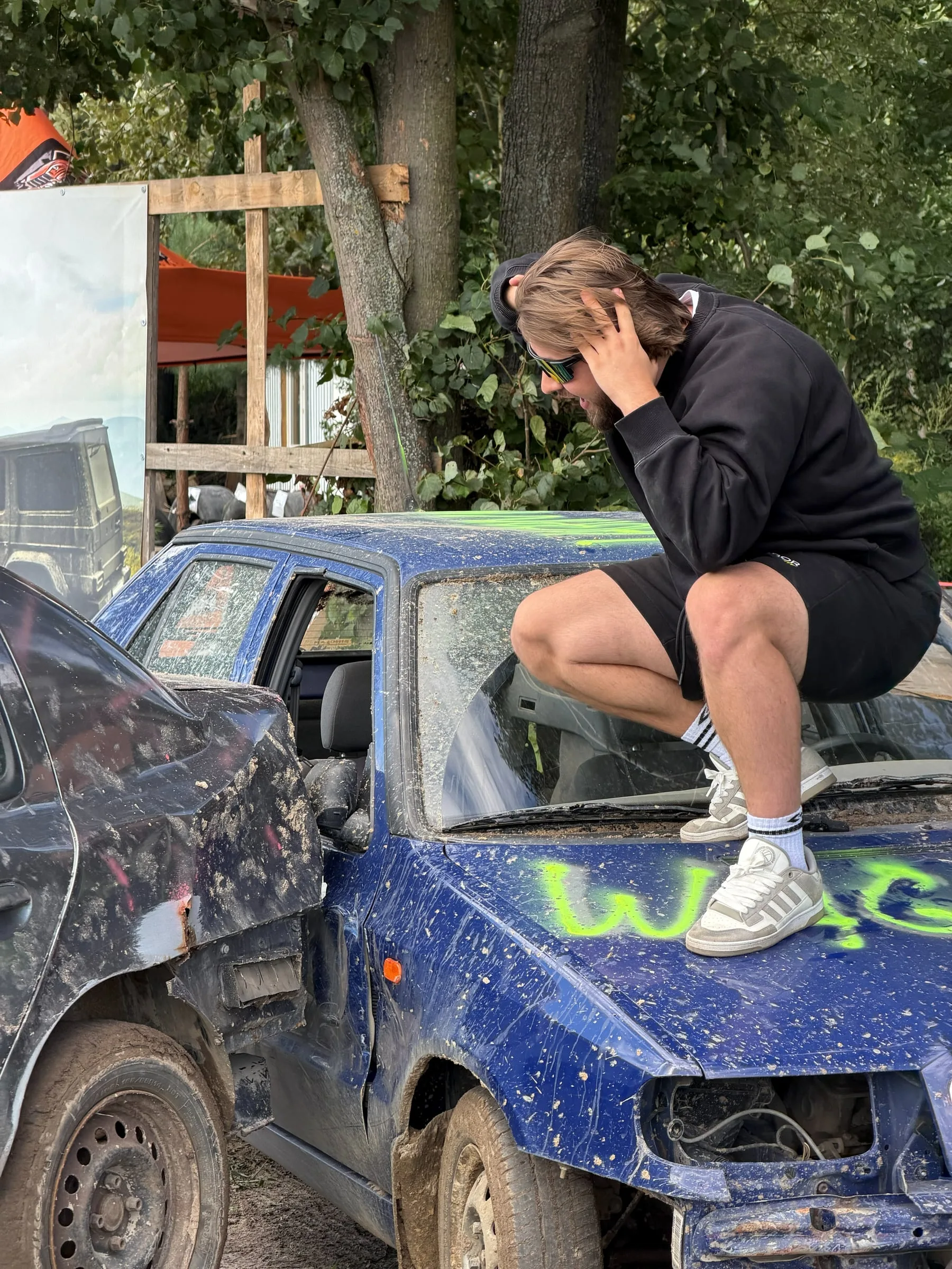 Participant dressed in black crouching on the bonnet of a battered blue car, hands on head