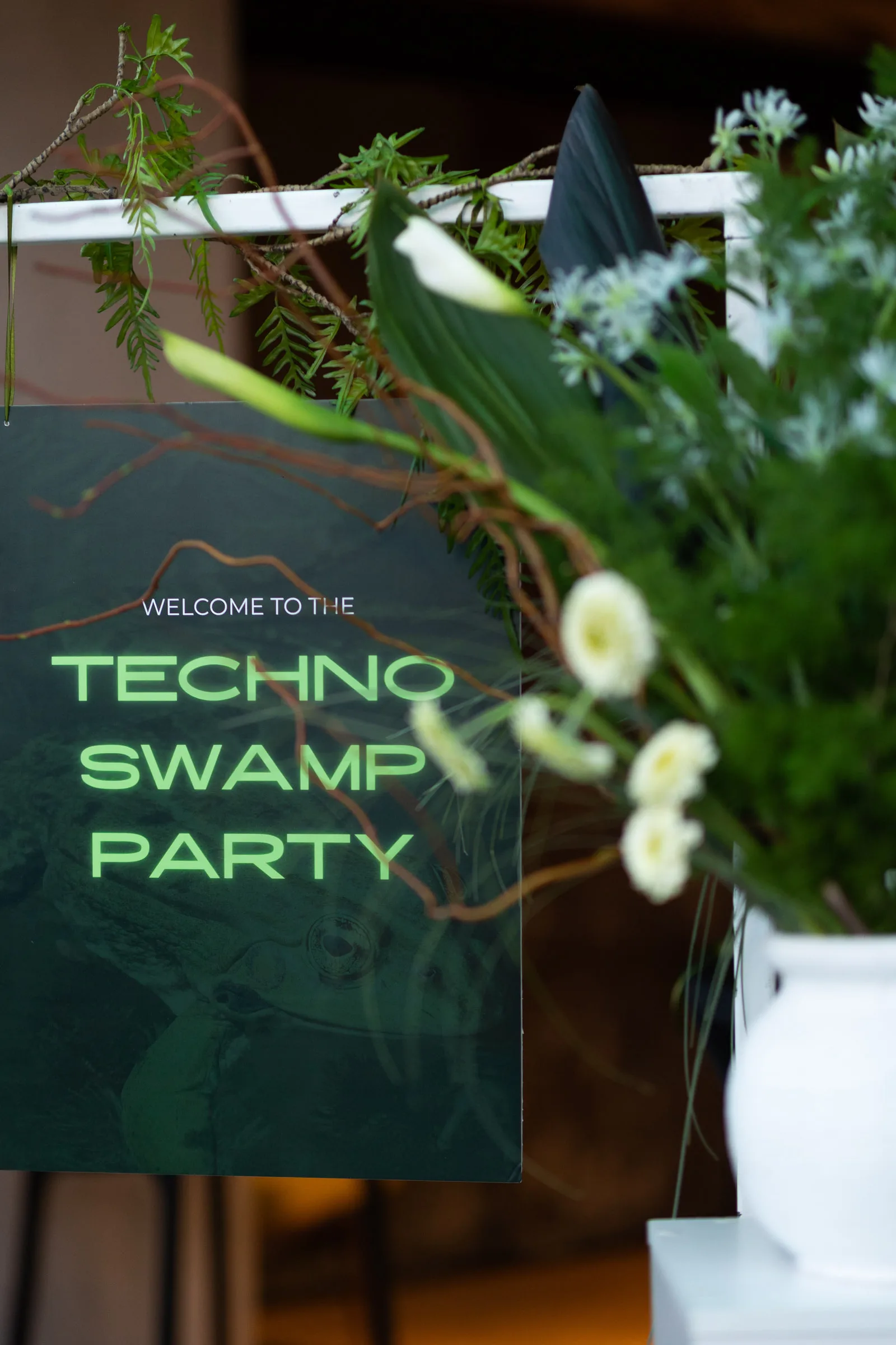 Табличка «Welcome to the Techno Swamp Party» серед зелені, гілок і білих квітів