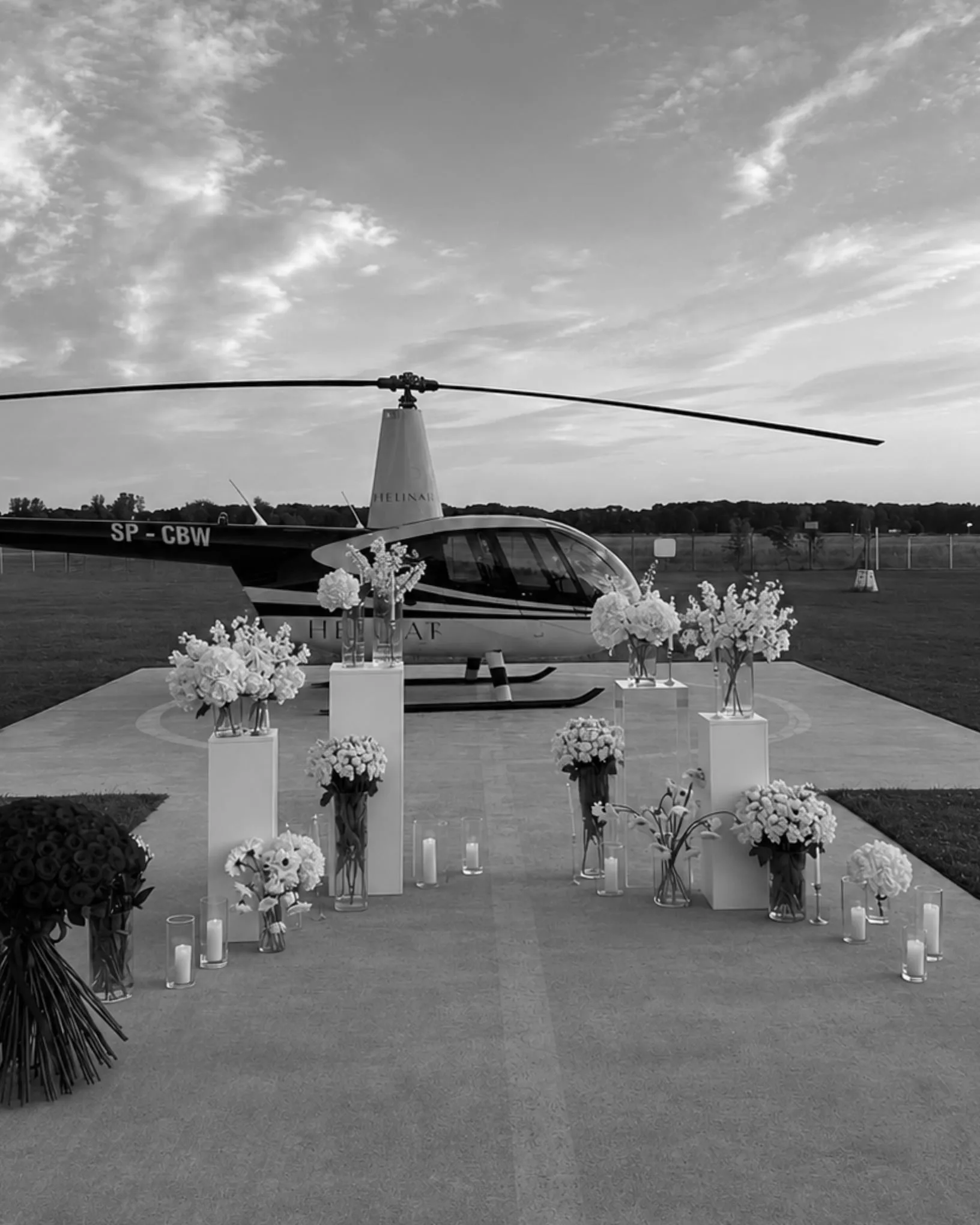 Black-and-white photo: white flowers in glass vases on white plinths and candles in glass holders in front of a helicopter