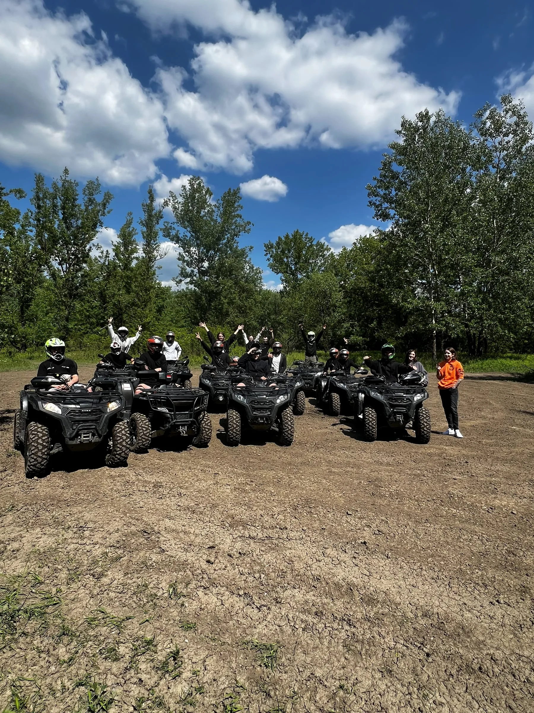 Participants in helmets on quad bikes raising their hands on a dirt clearing by the forest