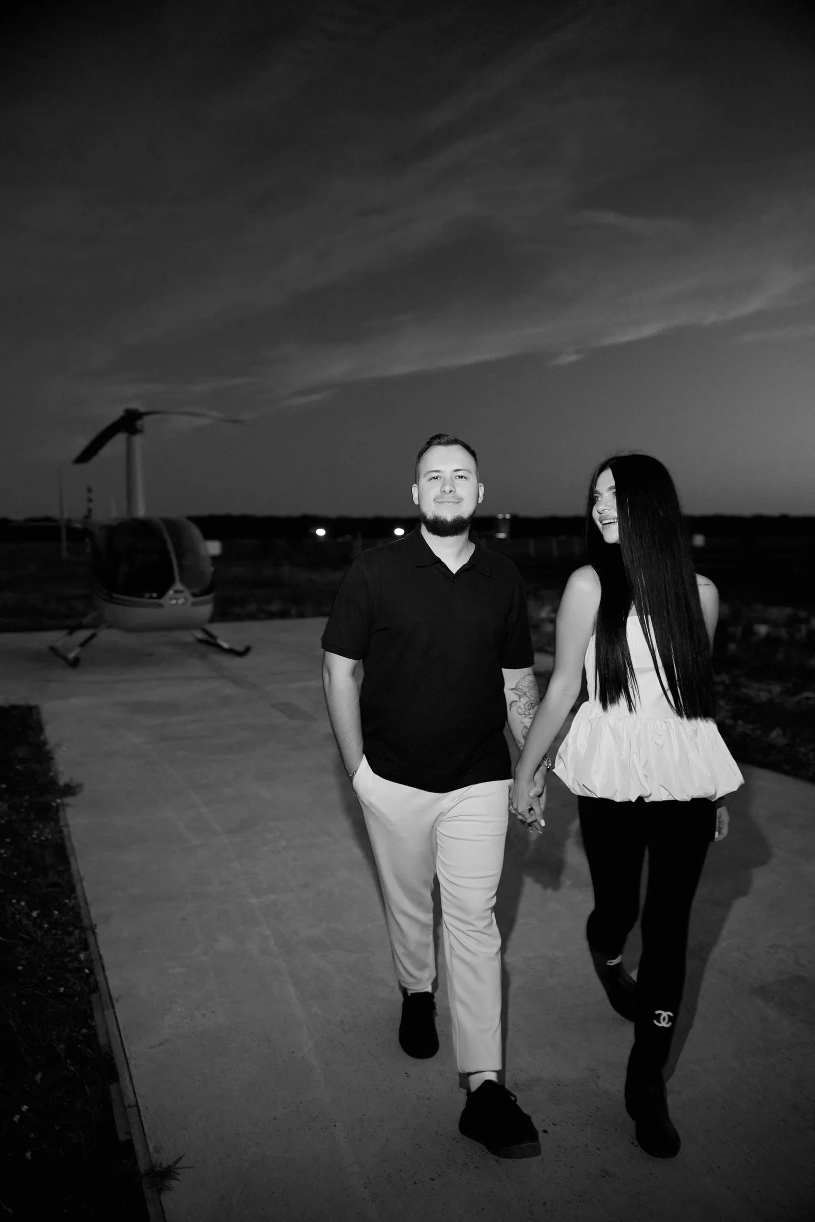 Black-and-white photo: a couple walking hand in hand across a helipad, with a helicopter behind them
