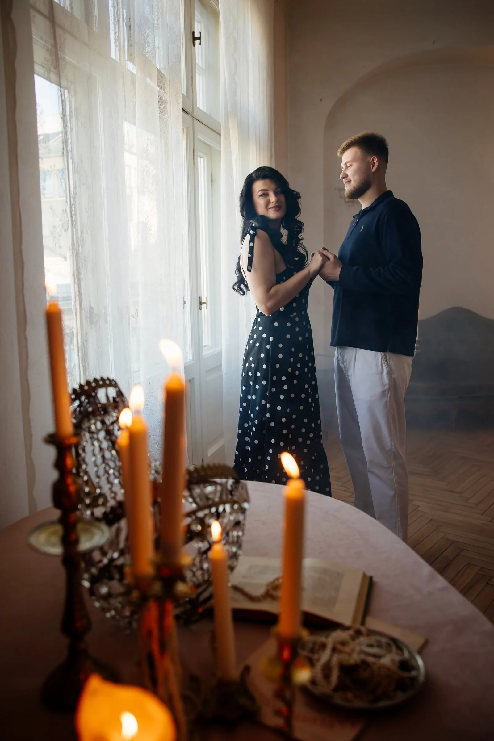 Чоловік і жінка тримаються за руки біля вікна, на передньому плані стіл зі свічками в канделябрах