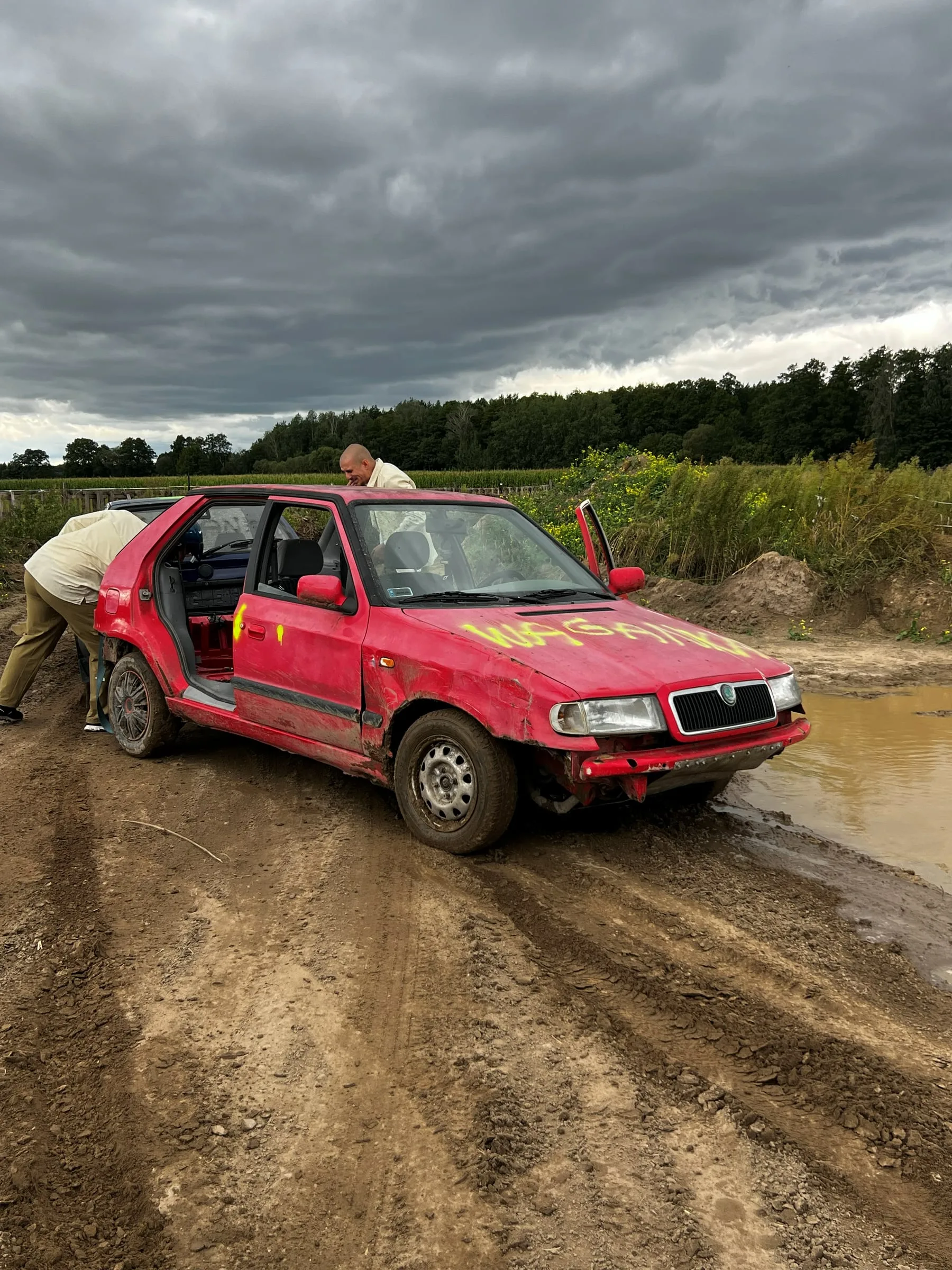 Two men beside a dented red car with spray-painted lettering on the bonnet, on a muddy track next to a puddle