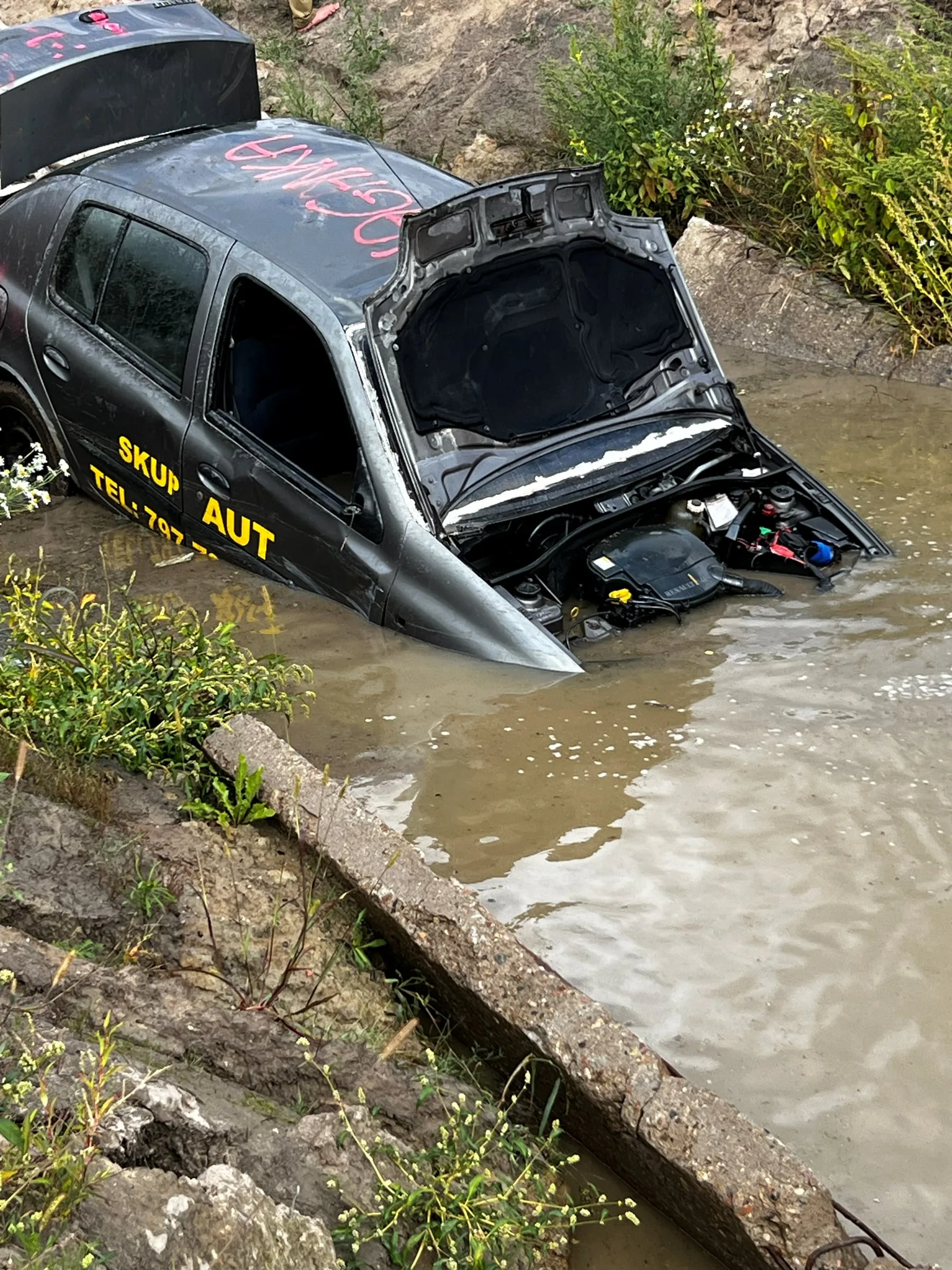 Dark, battered car with its bonnet open, half submerged in water