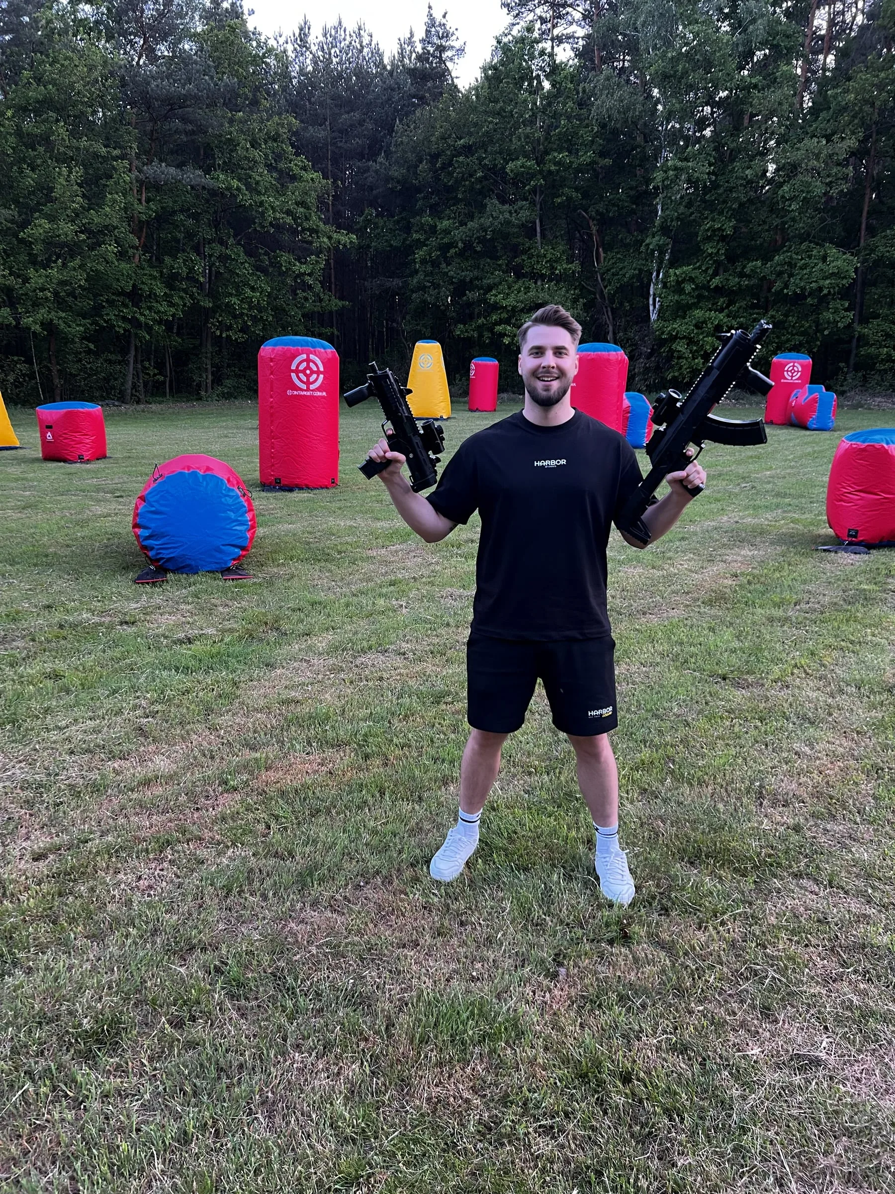 A Harbor team member in a branded T-shirt holding two game blasters on a field with inflatable obstacles
