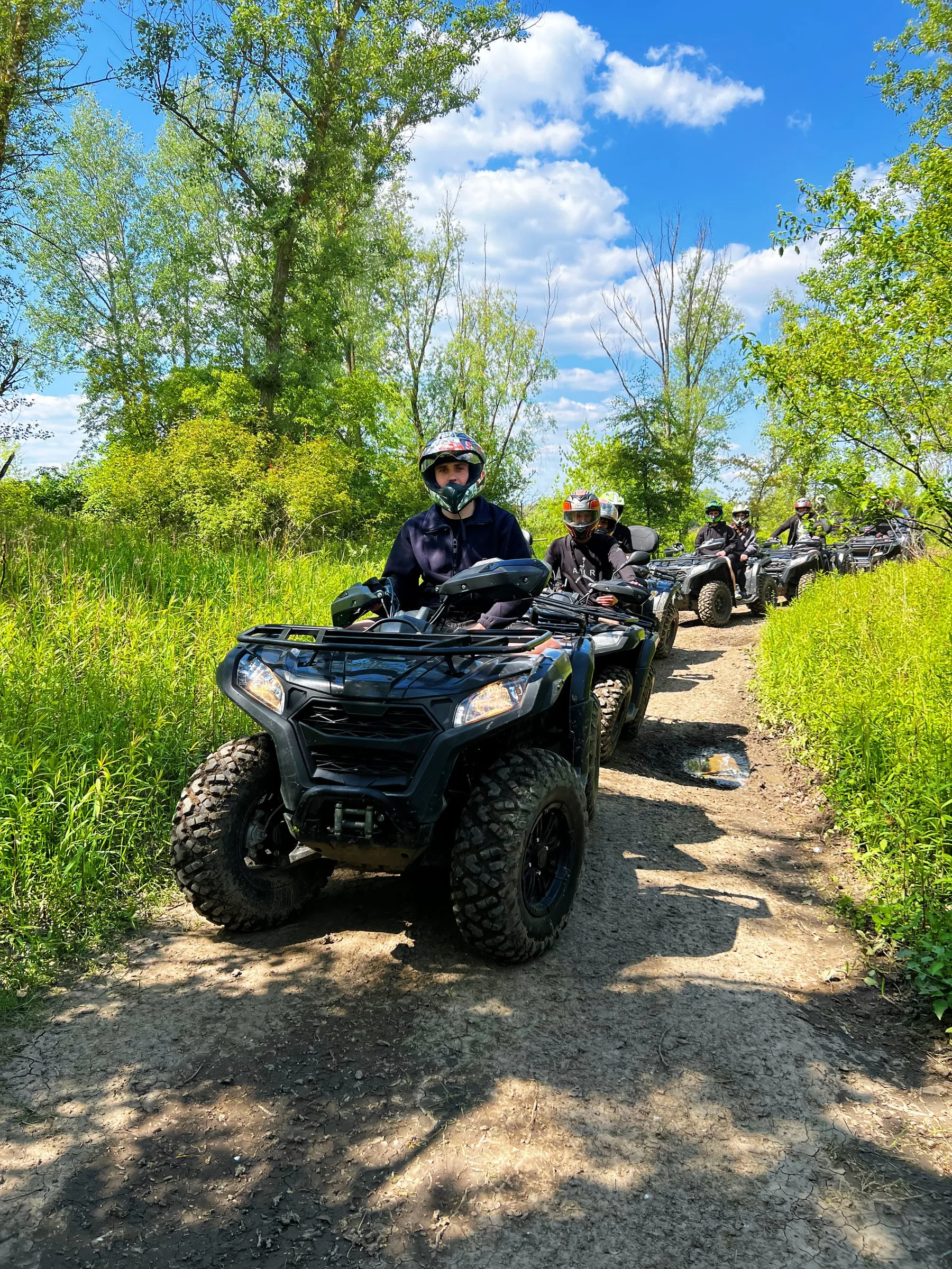 A line of participants in helmets riding quad bikes along a dirt trail through greenery