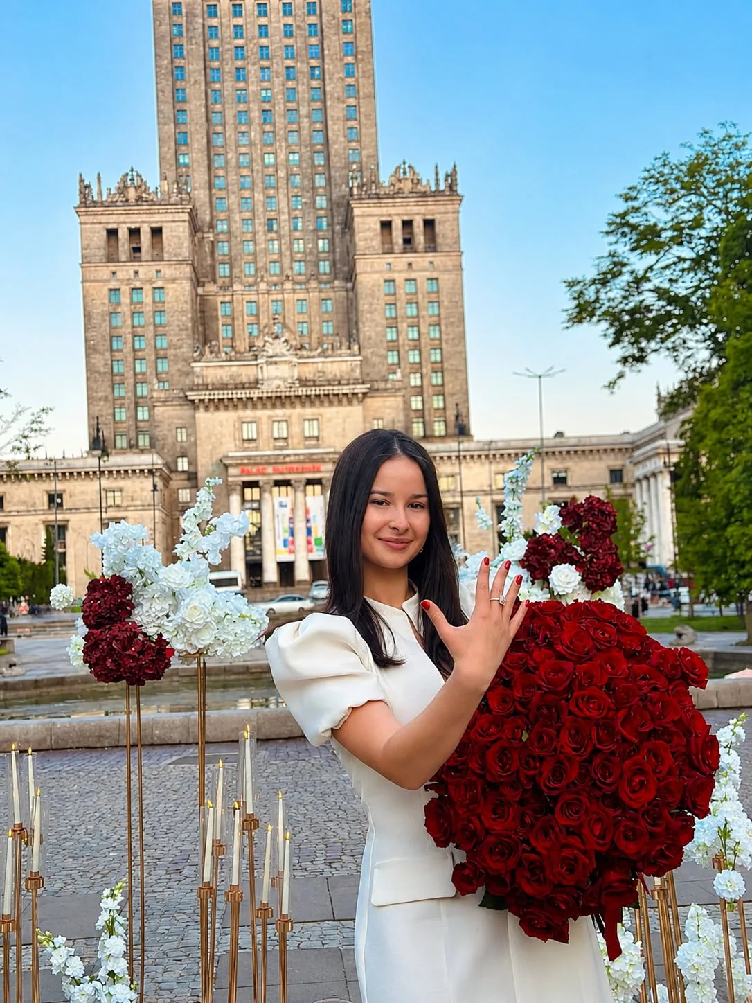 Dziewczyna z dużym bukietem czerwonych róż pokazuje pierścionek na tle Pałacu Kultury i Nauki