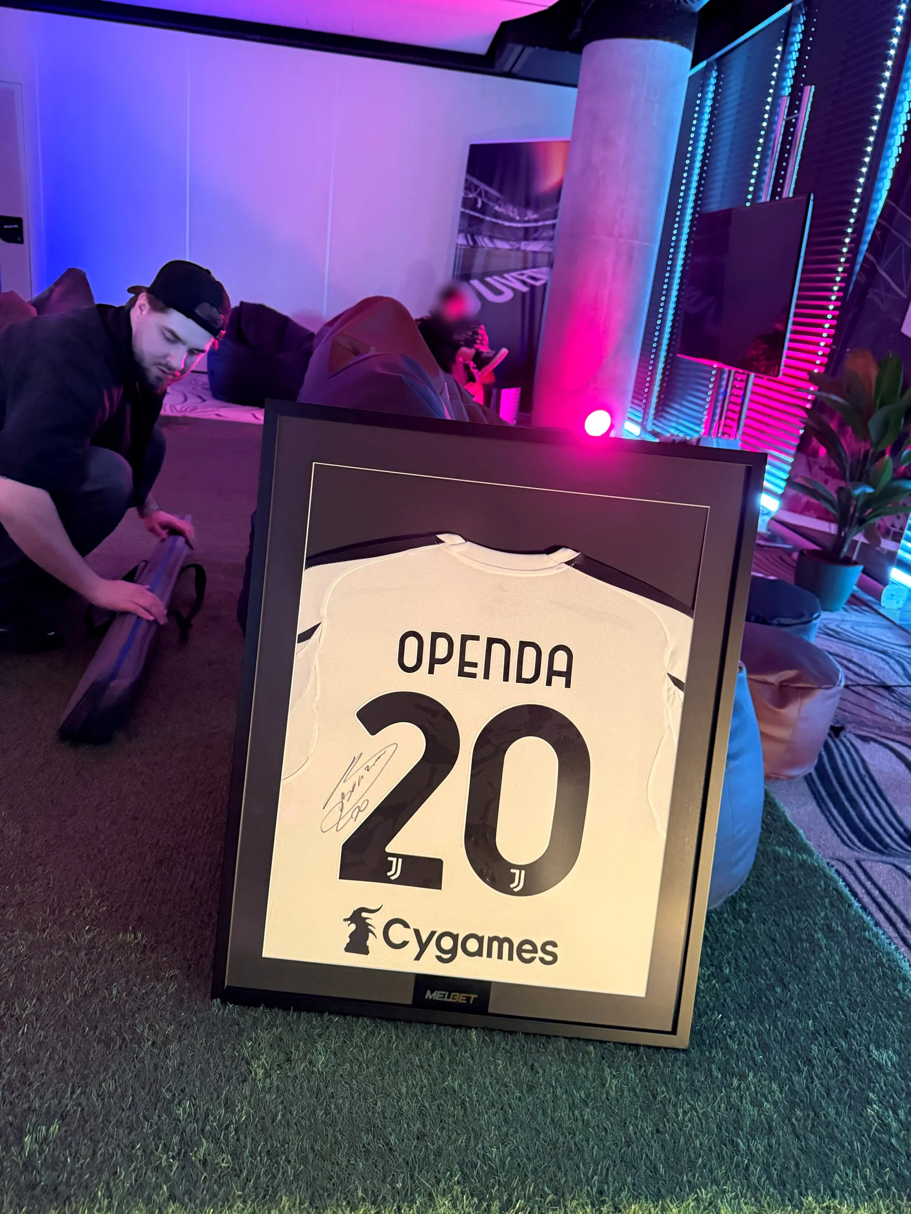 Mężczyzna kuca w strefie turniejowej obok ramy z podpisaną koszulką OPENDA, w tle miękkie pufy i neonowe podświetlenie