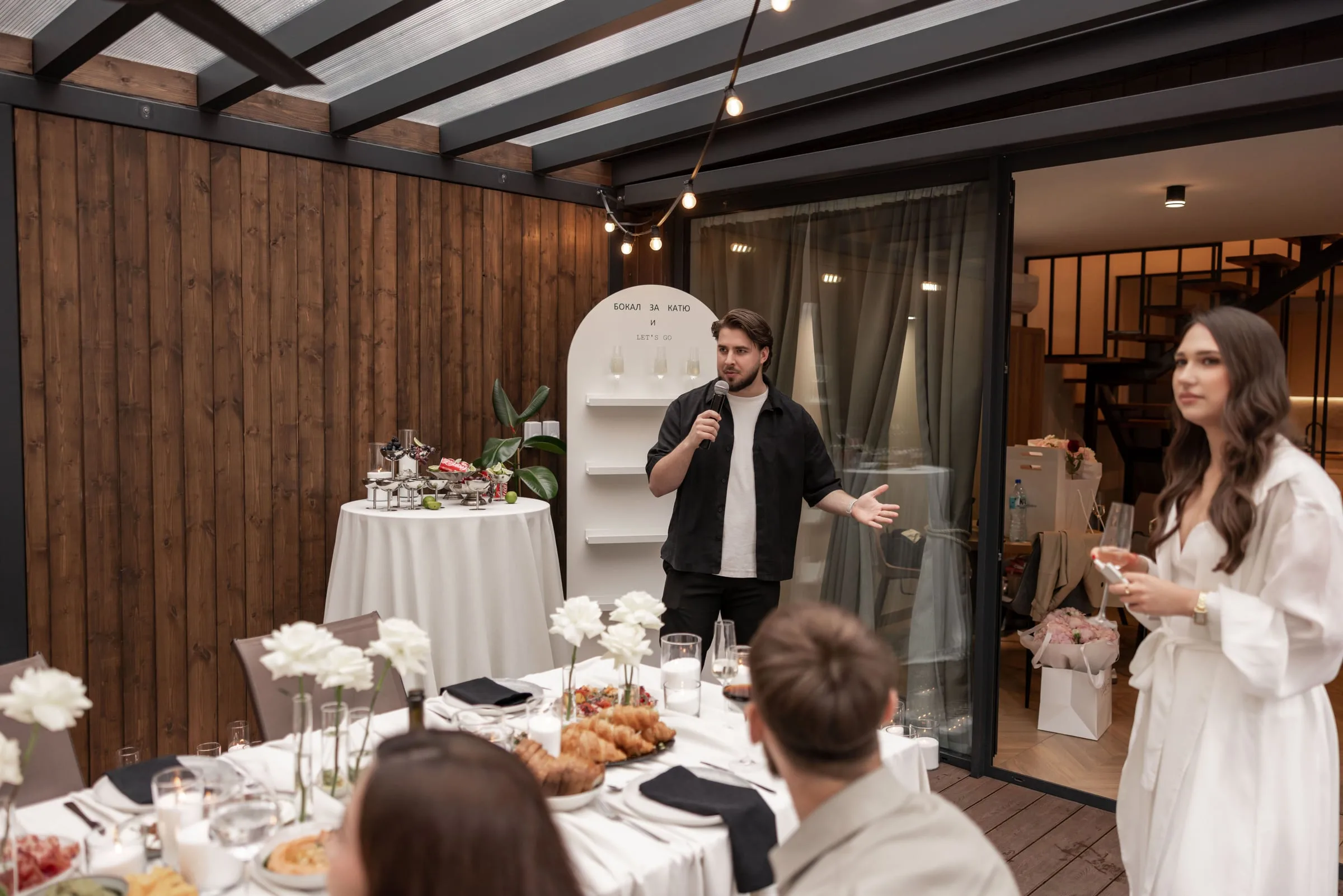 Host Makar speaking into a microphone to guests at a dinner table on a covered terrace