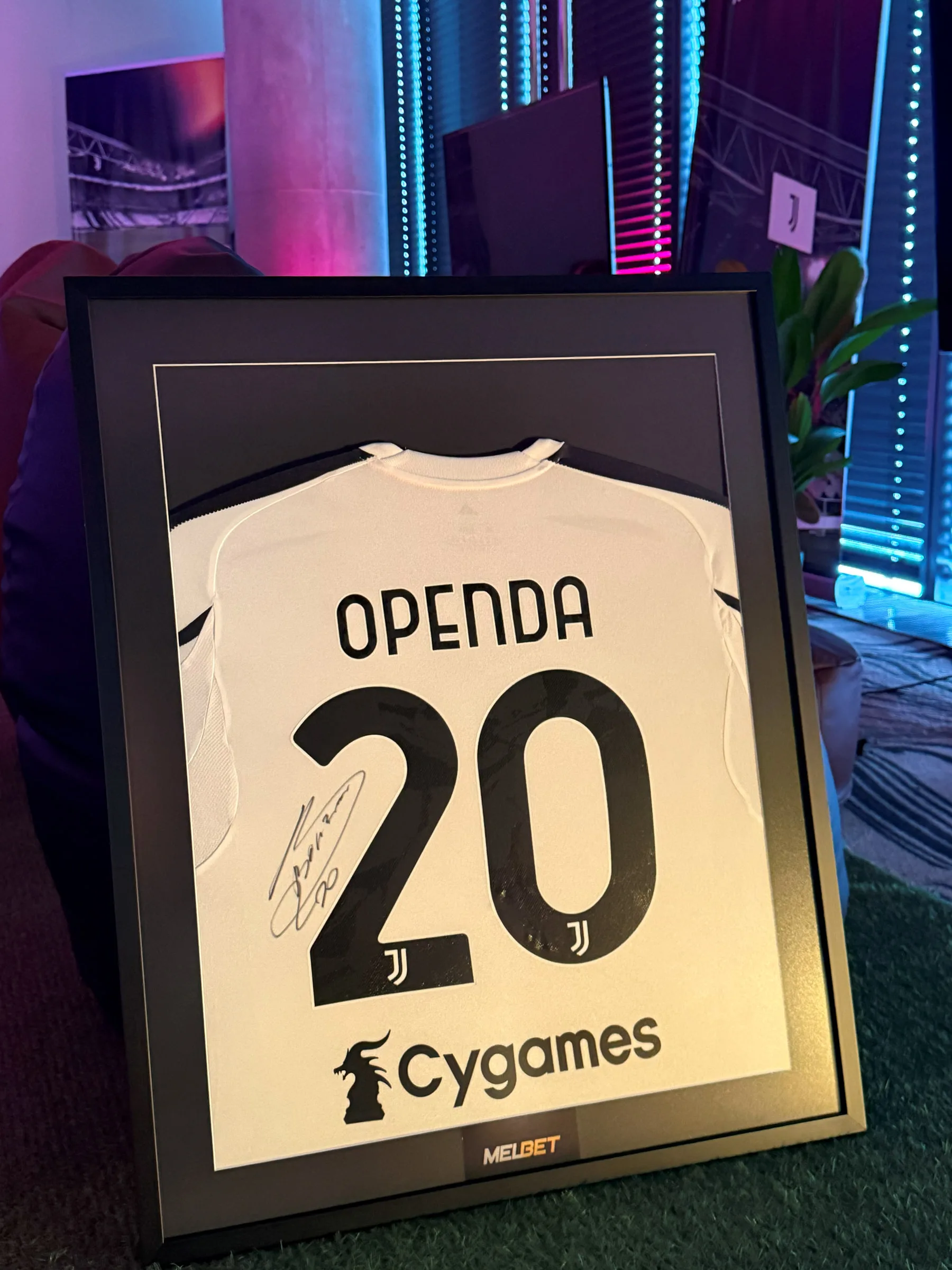 Rama z podpisaną koszulką Juventusu OPENDA z numerem 20 i tabliczką MELBET na tle neonowego podświetlenia