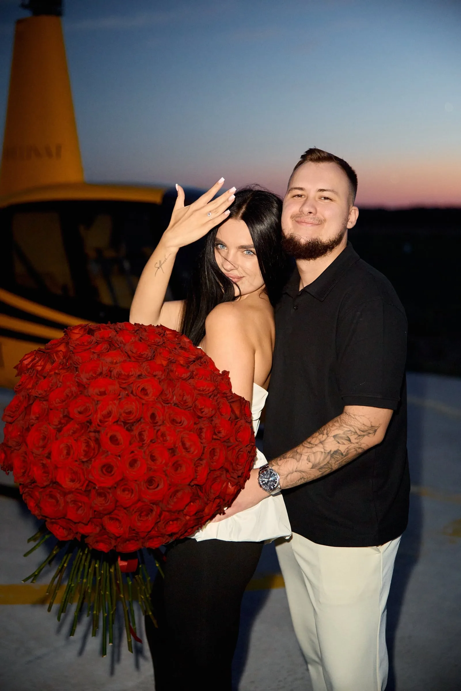A woman with a large bouquet of red roses shows her ring, with a smiling man beside her and a yellow helicopter behind
